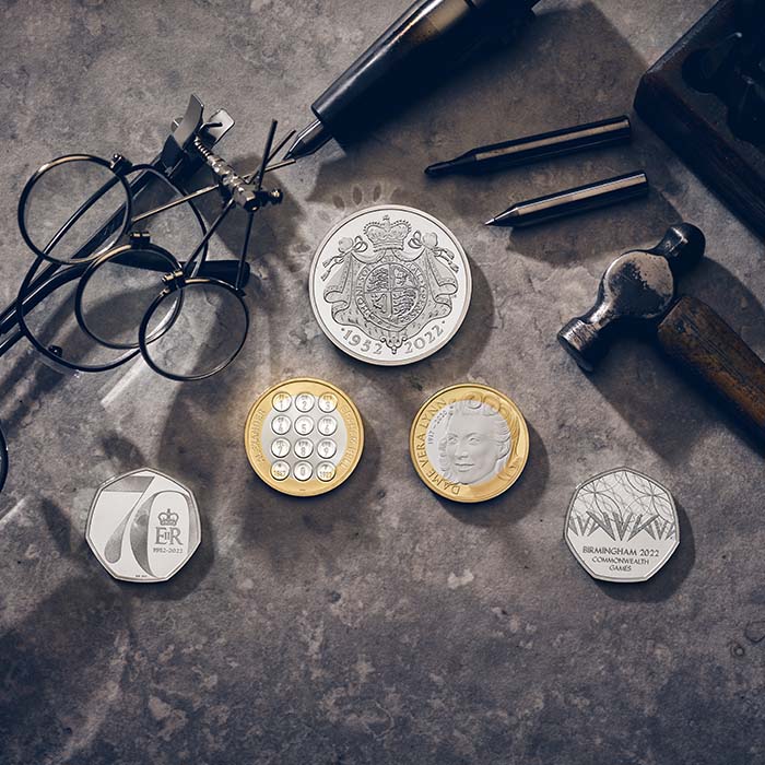 Collection of British coins produced by The Royal Mint on a textured surface with glasses, pen, and hammer